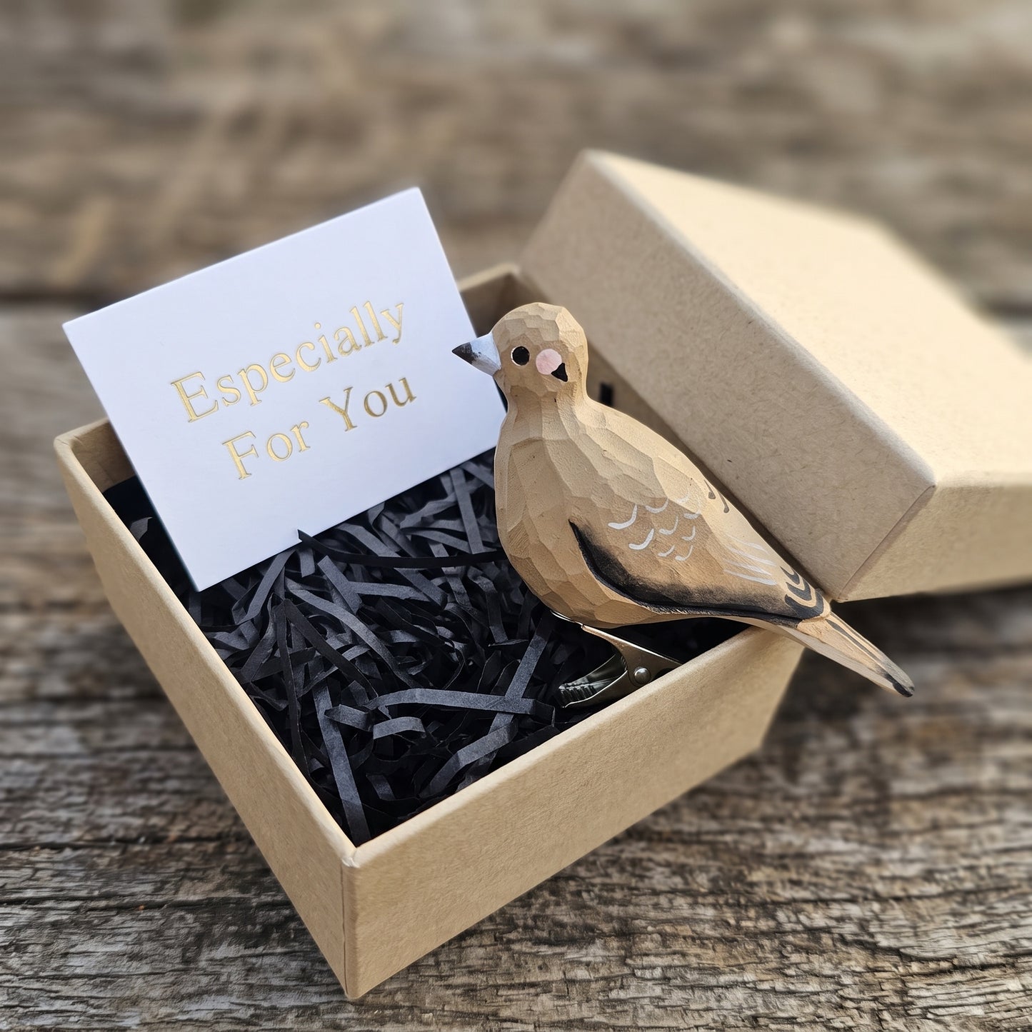 A hand-carved wooden mourning dove ornament resting inside an open kraft gift box filled with black shredded paper, accompanied by a white card with 'Especially For You' written in gold foil.