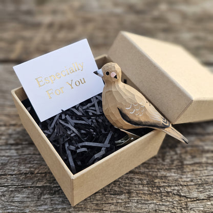 A hand-carved wooden mourning dove ornament resting inside an open kraft gift box filled with black shredded paper, accompanied by a white card with 'Especially For You' written in gold foil.