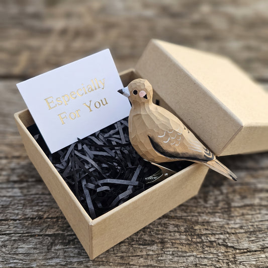A hand-carved wooden mourning dove ornament resting inside an open kraft gift box filled with black shredded paper, accompanied by a white card with 'Especially For You' written in gold foil.