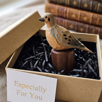 A carved wooden Mourning Dove figurine displayed inside a kraft gift box with black shredded paper filler and a card that reads 'Especially For You.' The bird has a light natural wood body with dark eye and beak details, spotted wing patterns, and a small blue accent on its back, mounted on a dark wooden base.