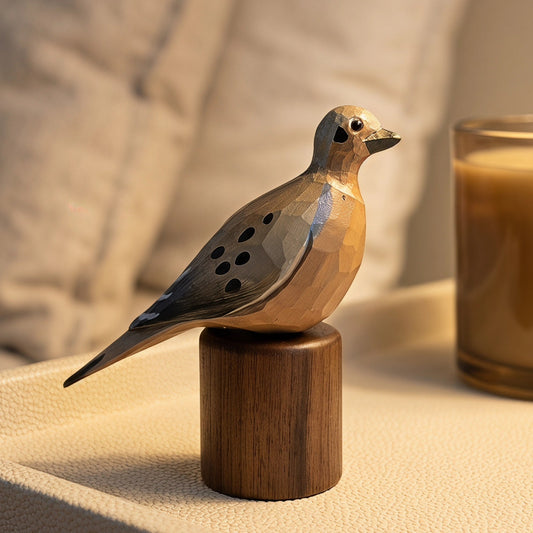 A small geometric wooden Mourning Dove figurine with spotted detailing stands on a cylindrical wooden base on a textured cream surface, with a warm-toned candle and soft pillows blurred in the background.