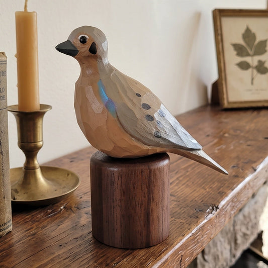 A hand-painted wooden Mourning Dove figurine on a dark wooden cylindrical base, displayed on a rustic wooden mantel next to vintage books, a candle, and a framed botanical print.