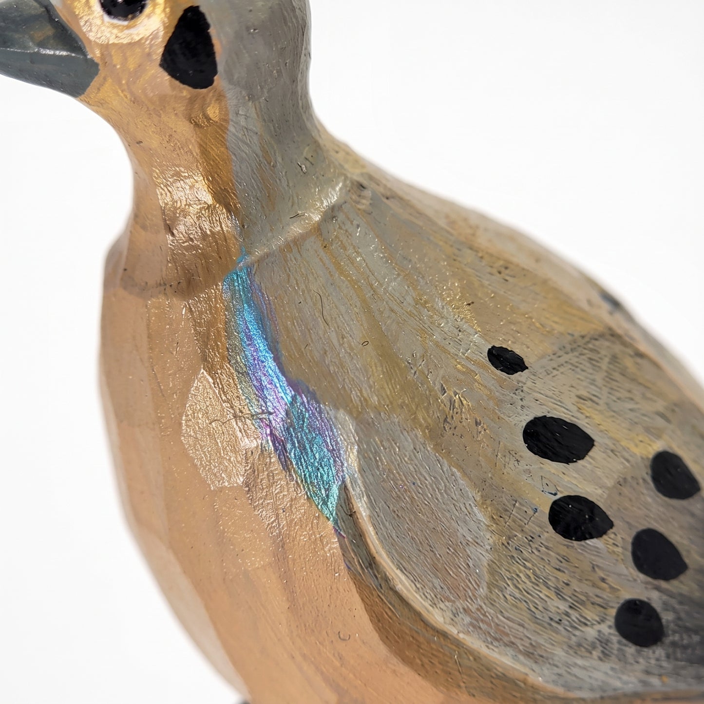 A close-up of a handcrafted wooden Mourning Dove figurine, showing its textured natural wood grain, black painted spots, and a small iridescent blue-green accent on its back against a white background.