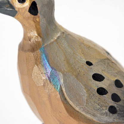 A close-up of a handcrafted wooden Mourning Dove figurine, showing its textured natural wood grain, black painted spots, and a small iridescent blue-green accent on its back against a white background.
