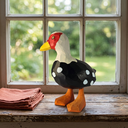 Hand-carved wooden Muscovy Duck figurine standing on a rustic wooden windowsill beside a folded terracotta cloth, with a sunny garden visible through the window.