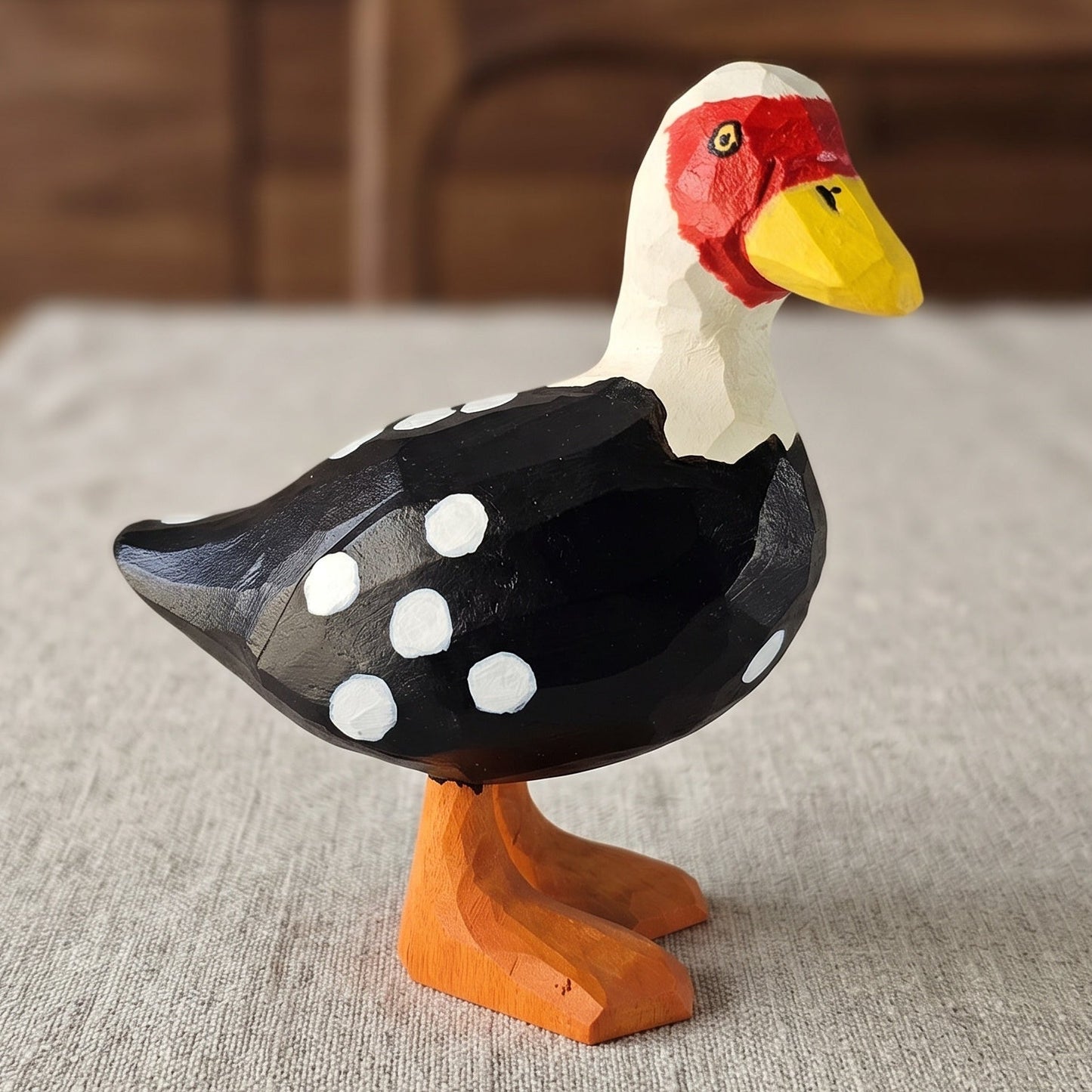 Hand-carved wooden Muscovy Duck figurine with black and white dotted plumage, bright red caruncled face, yellow bill, and orange webbed feet resting on a linen tablecloth.