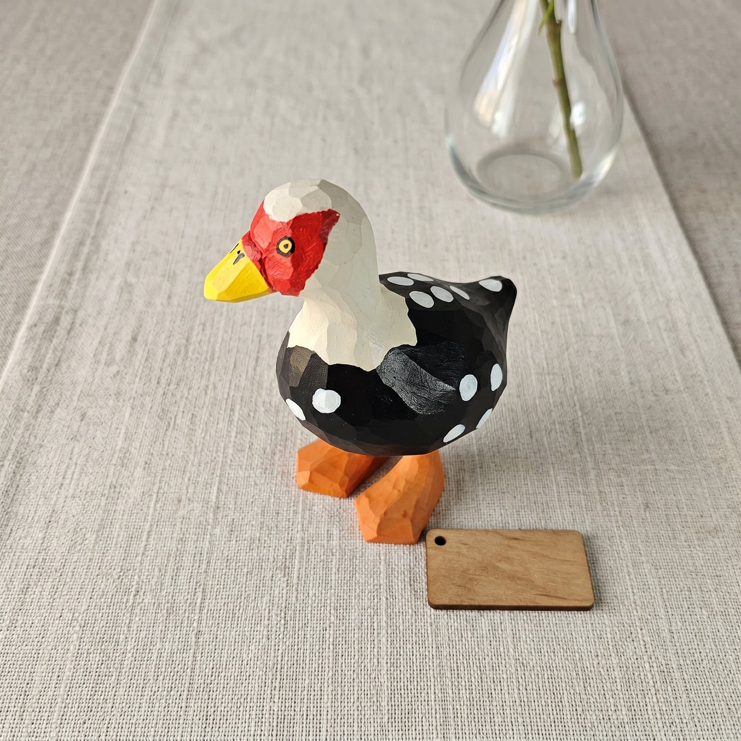 Hand-carved wooden Muscovy Duck figurine with black and white dotted plumage, red face mask, and orange feet displayed next to a blank wooden tag on a linen table runner.