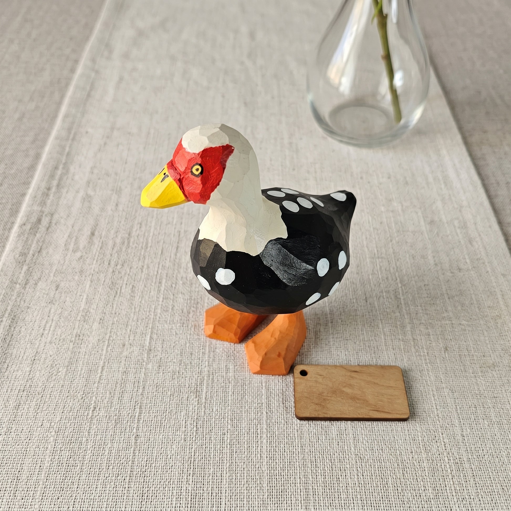 Hand-carved wooden Muscovy Duck figurine with black and white dotted plumage, red face mask, and orange feet displayed next to a blank wooden tag on a linen table runner.
