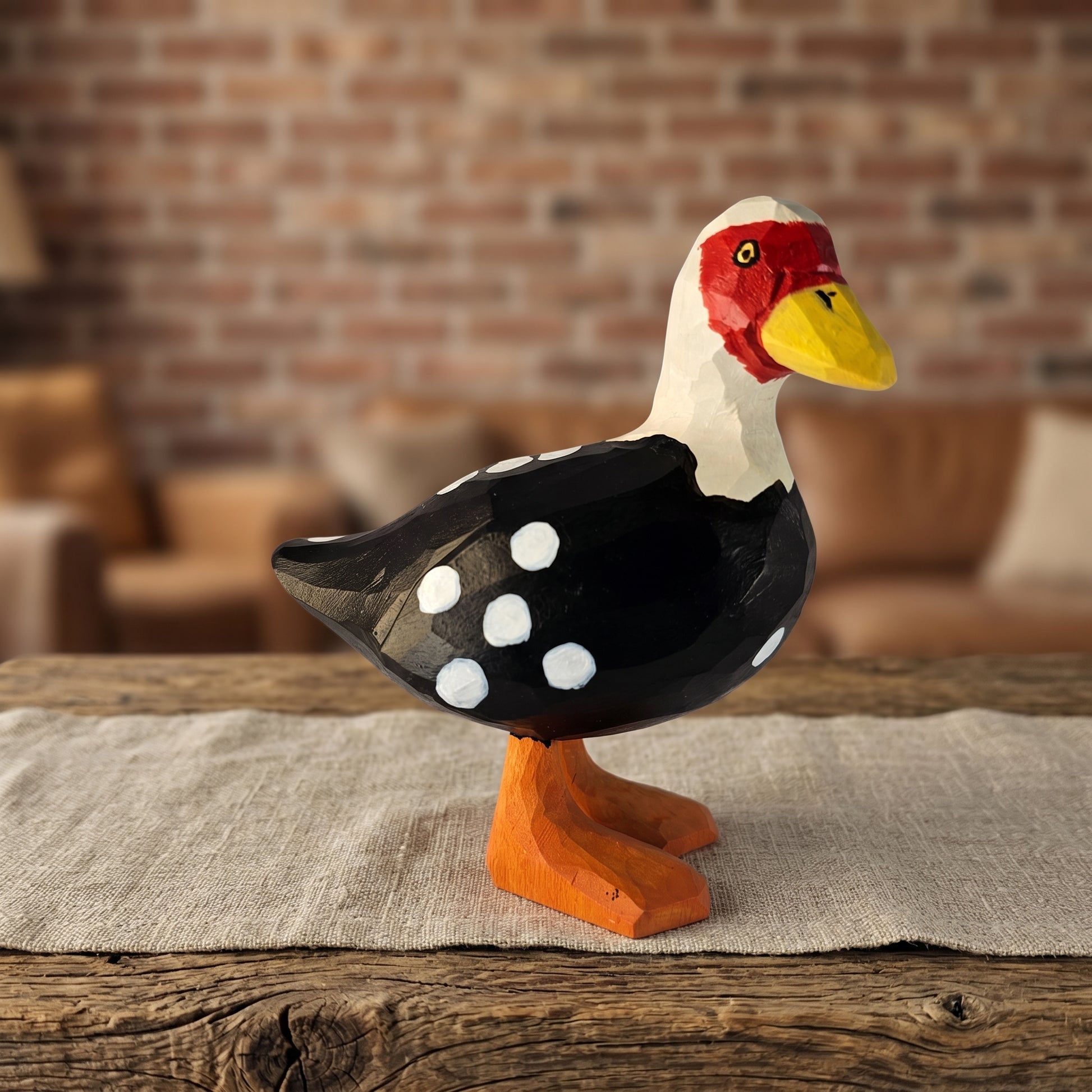 Hand-carved wooden Muscovy Duck figurine with black and white spotted wings, red facial mask, yellow bill, and orange webbed feet standing on a rustic wooden table in a warm room setting.