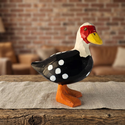 Hand-carved wooden Muscovy Duck figurine with black and white spotted wings, red facial mask, yellow bill, and orange webbed feet standing on a rustic wooden table in a warm room setting.