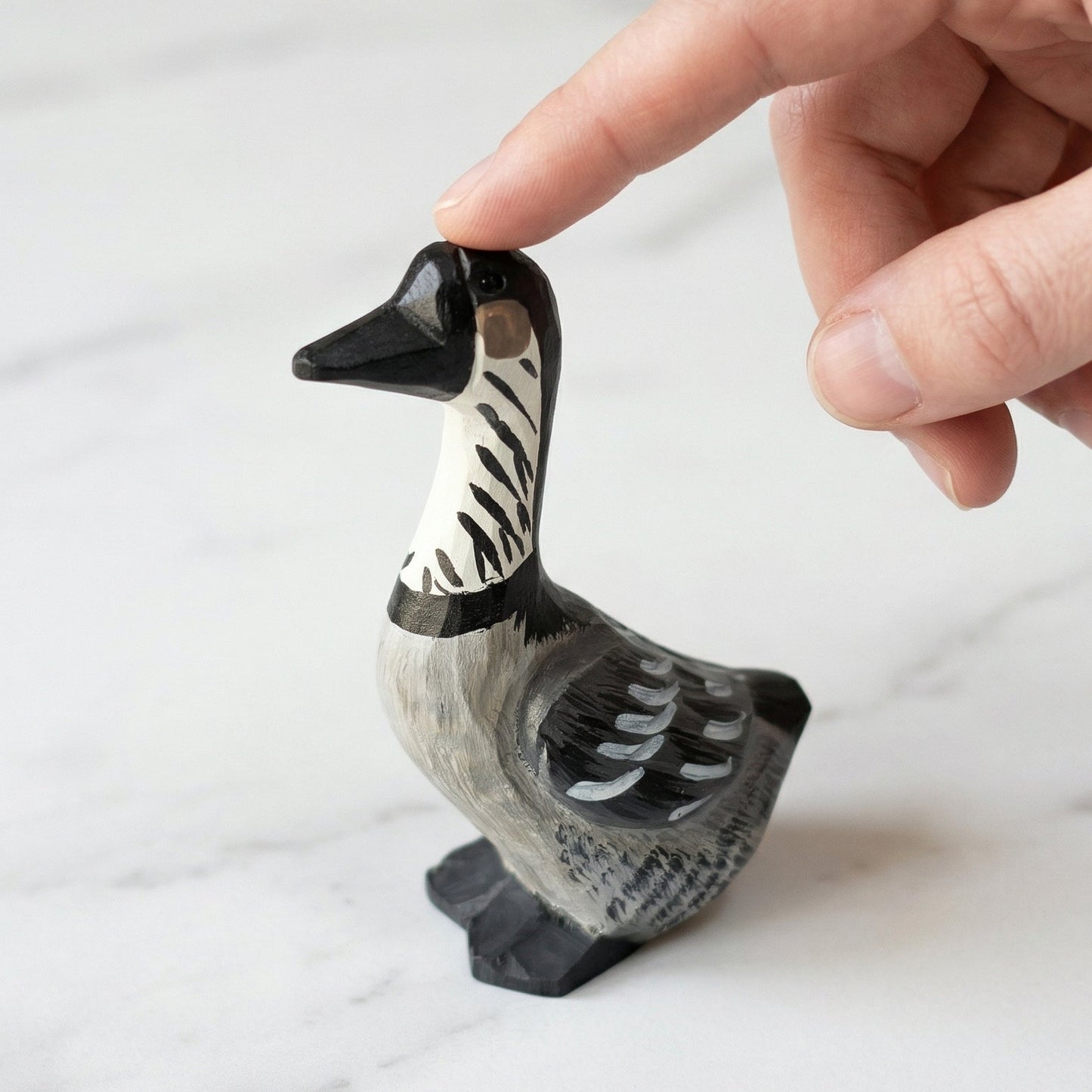 A hand is gently touching the top of a small carved duck figurine. The figurine has a black and white patterned neck and a gray and black painted body, standing on a light marble surface.
