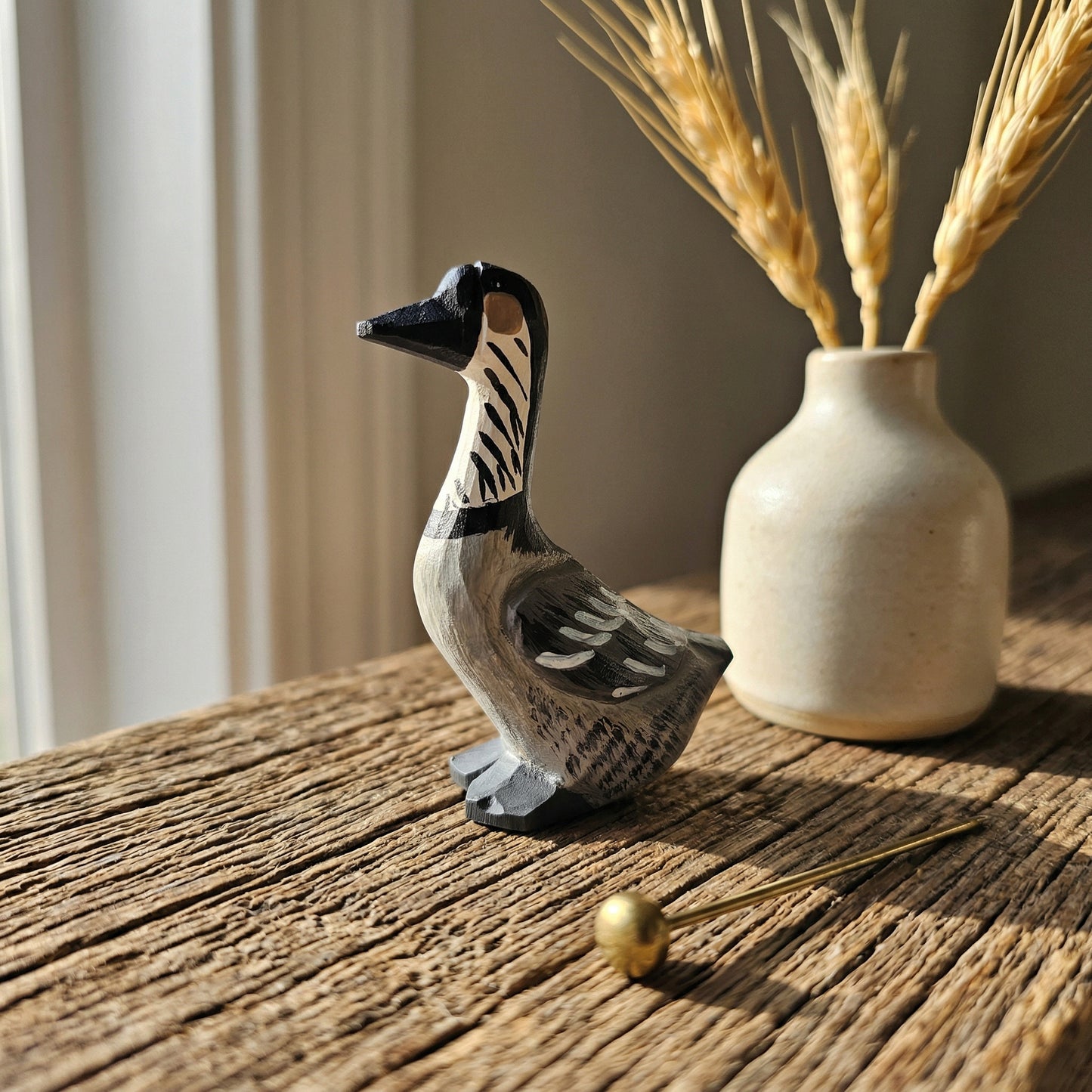 A hand-painted wooden Hawaiian Goose figurine with black, white, and gray details, displayed on a rustic wooden table next to a small ceramic vase with dried wheat stalks.