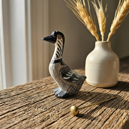 A hand-painted wooden Hawaiian Goose figurine with black, white, and gray details, displayed on a rustic wooden table next to a small ceramic vase with dried wheat stalks.