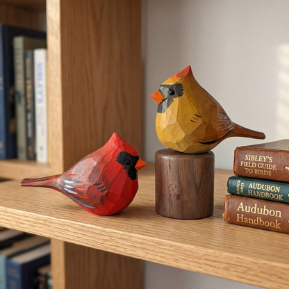 Two geometric, faceted wooden cardinal bird sculptures displayed on a wooden bookshelf next to a stack of vintage bird-handbooks.