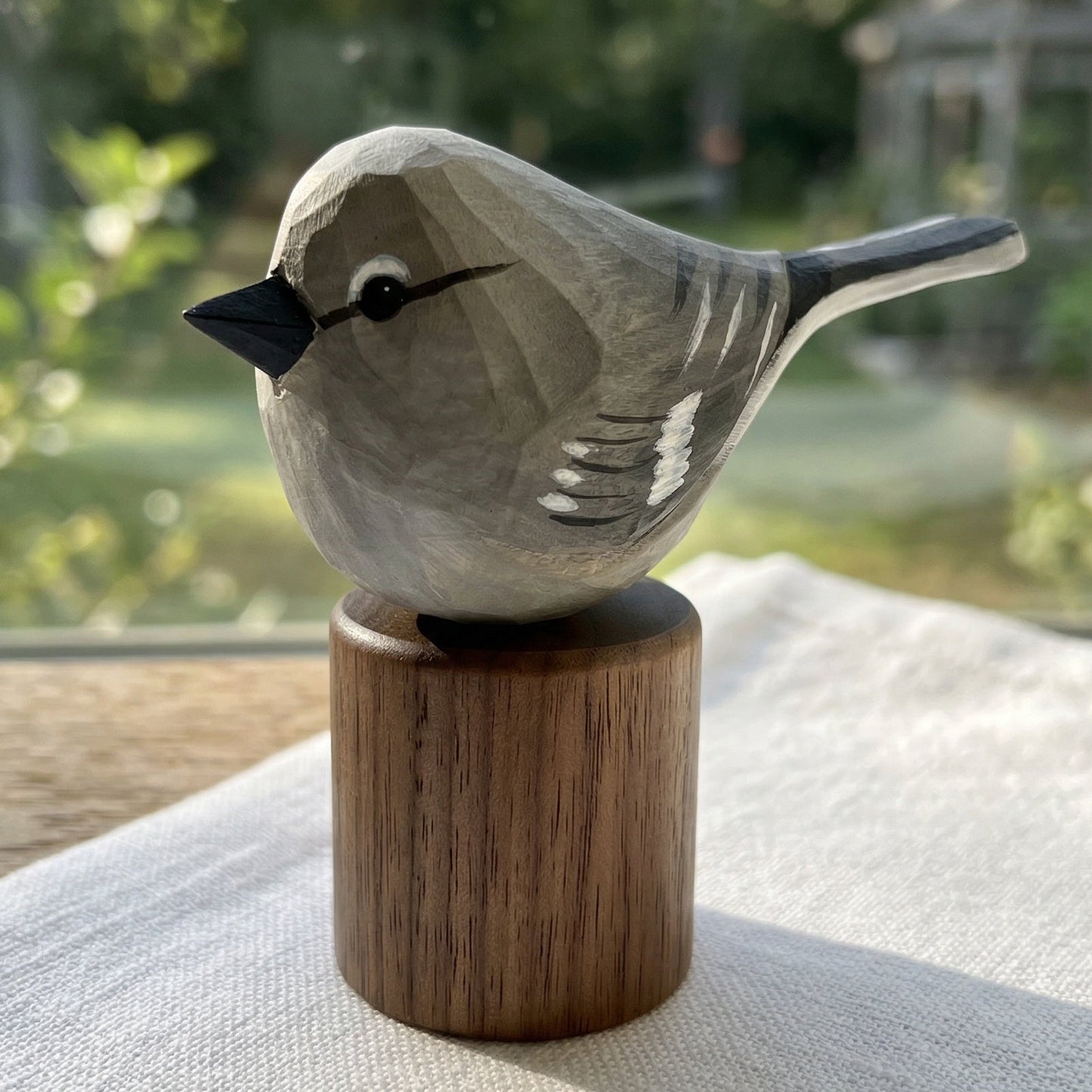 A hand-carved wooden Northern mockingbird figurine with a gray and white painted finish and a dark beak, mounted on a cylindrical wooden base. The figurine is displayed on a light-colored fabric surface with a blurred garden background visible through a window.