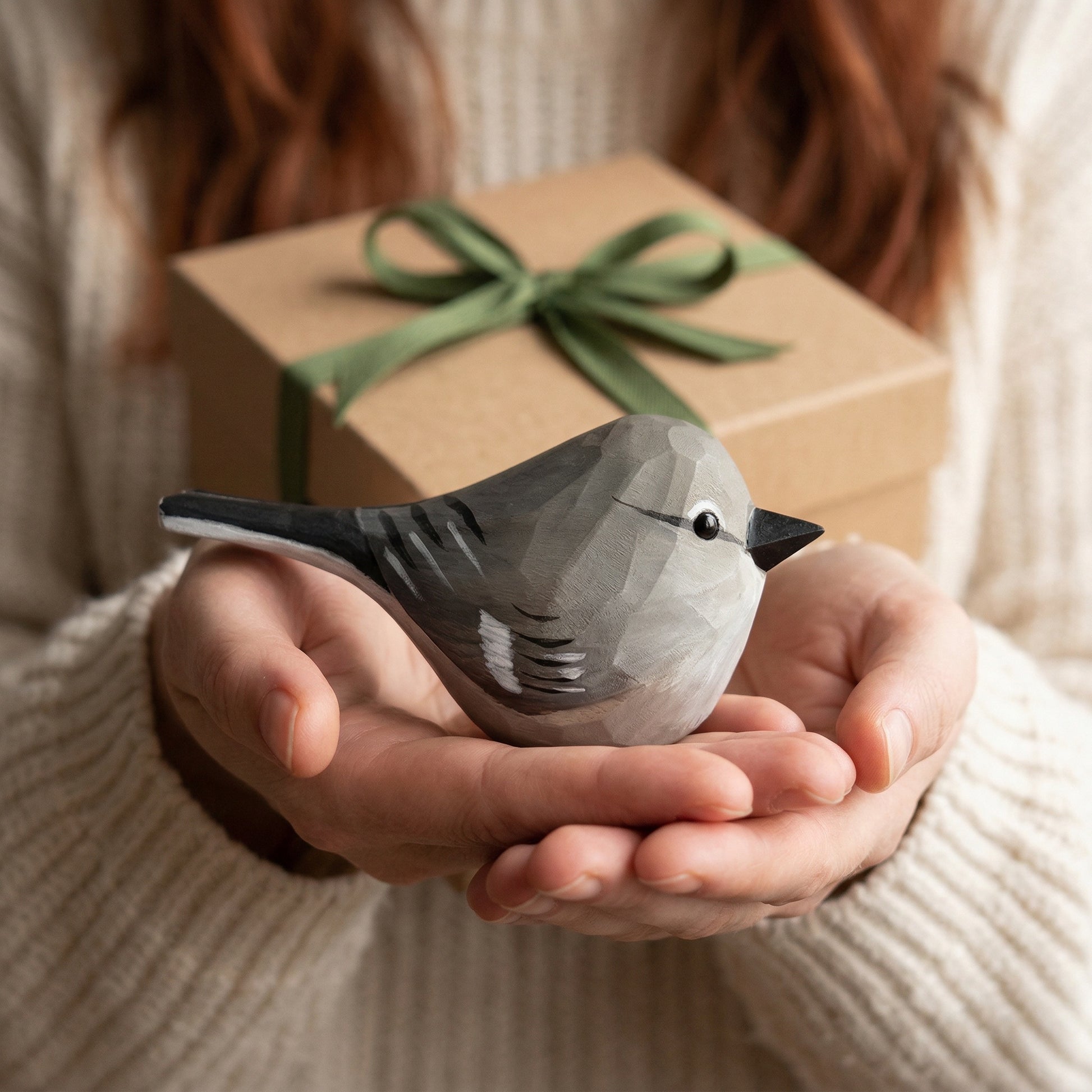 A person in a cream knit sweater holds a small gray Northern mockingbird figurine in their hands, with a brown gift box and green ribbon in the background.