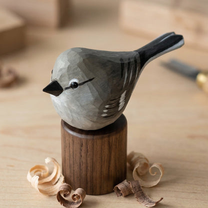 A hand-carved wooden Northern mockingbird figurine with a gray and black painted finish, mounted on a dark wood pedestal. The bird is set on a light wood surface with wood shavings scattered around it, and a blurred carving knife in the background.