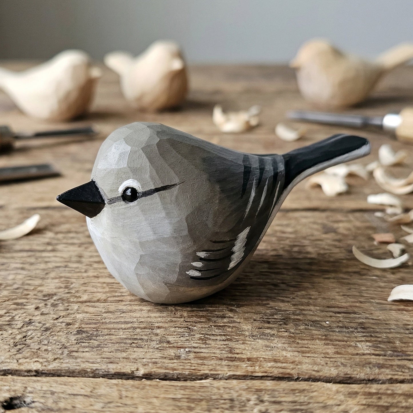 A hand-carved wooden Northern mockingbird figurine with a gray and black painted finish, resting on a rustic wooden workbench surrounded by wood shavings and carving tools.