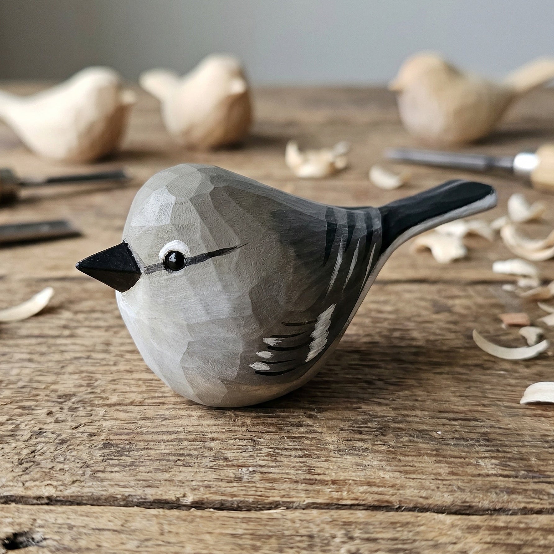 A hand-carved wooden Northern mockingbird figurine with a gray and black painted finish, resting on a rustic wooden workbench surrounded by wood shavings and carving tools.
