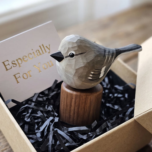 A small carved Northern mockingbird figurine with a geometric, faceted design sits on a wooden stand inside a kraft gift box filled with black shredded paper and a card that reads 'Especially For You'.