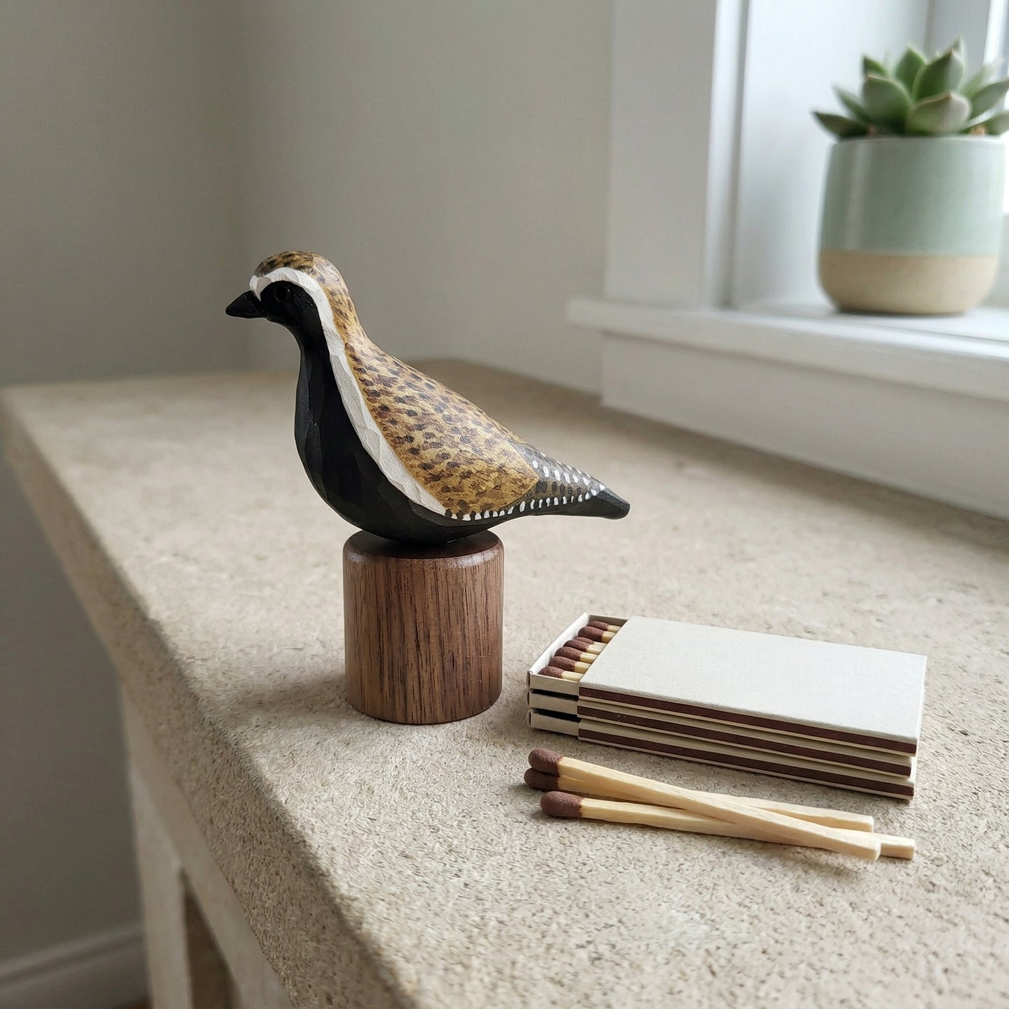 Hand-carved wooden Pacific Golden Plover figurine on a cylindrical black walnut base resting on a stone ledge next to matchsticks, a notepad, and a potted succulent.