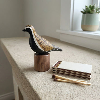Hand-carved wooden Pacific Golden Plover figurine on a cylindrical black walnut base resting on a stone ledge next to matchsticks, a notepad, and a potted succulent.