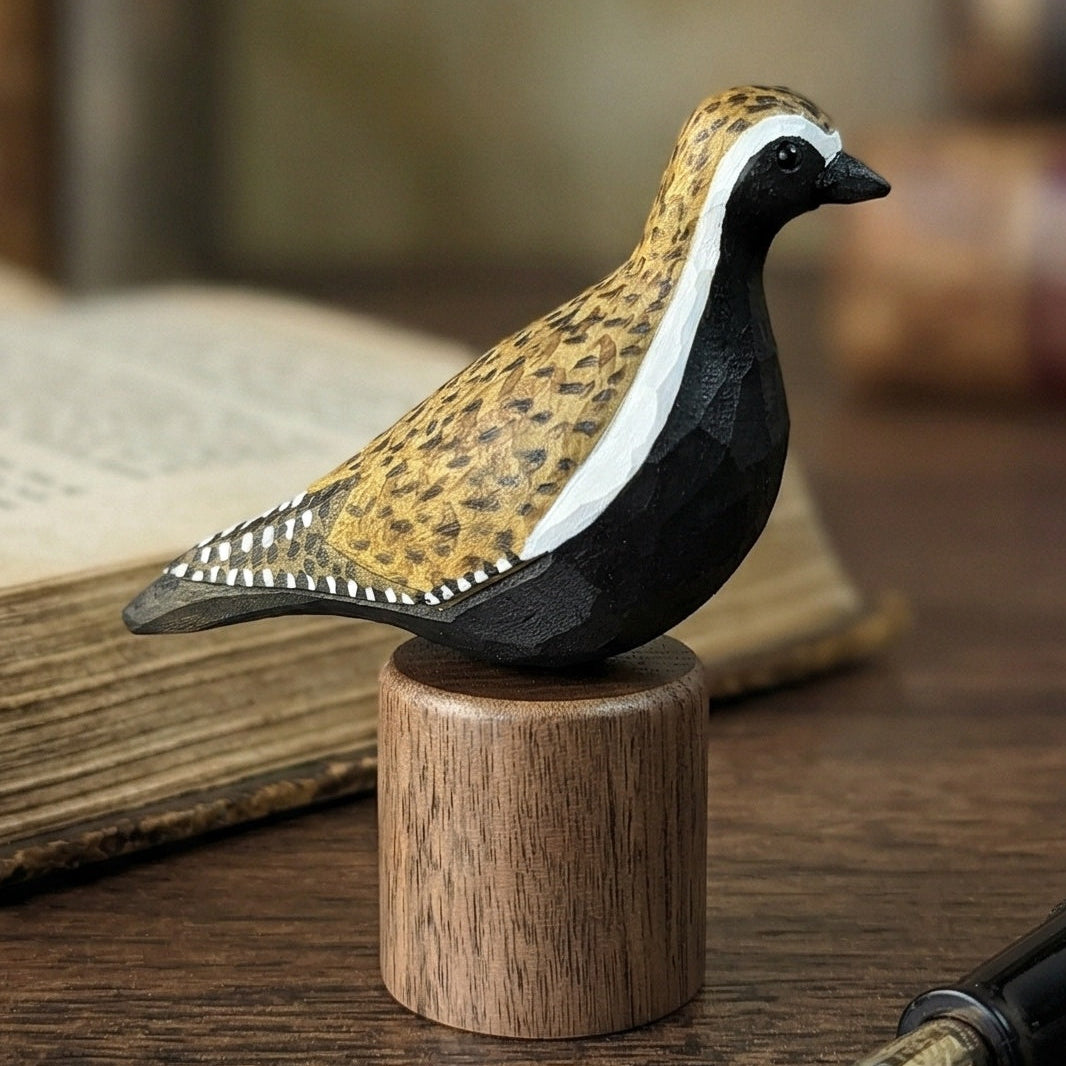 Side view of a hand-carved Pacific Golden Plover wooden bird figurine on a black walnut base next to a fountain pen and an open vintage book.