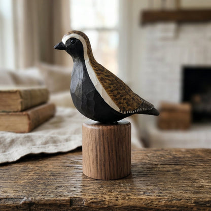 Hand-carved wooden Pacific Golden Plover bird figurine perched on a cylindrical black walnut base on a rustic wooden table with vintage books in the background.