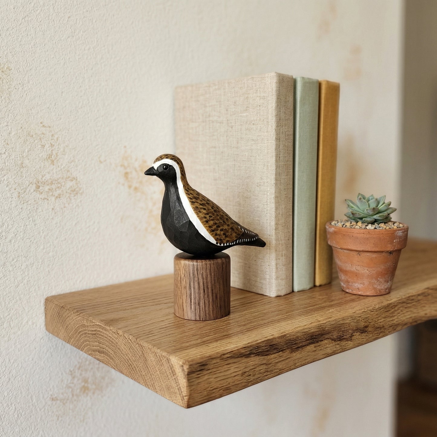 Hand-carved wooden Pacific Golden Plover bird figurine perched on a cylindrical wooden base, displayed on a floating wooden wall shelf next to books and a potted succulent.