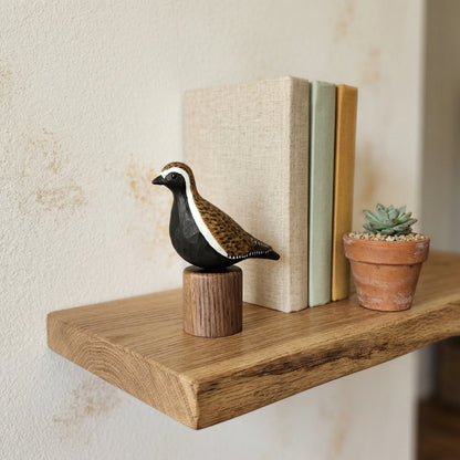 Hand-carved wooden Pacific Golden Plover bird figurine perched on a cylindrical wooden base, displayed on a floating wooden wall shelf next to books and a potted succulent.
