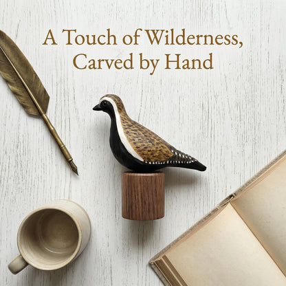 Promotional graphic with text "A Touch of Wilderness, Carved by Hand" featuring a hand-carved Pacific Golden Plover wooden bird figurine on a black walnut base, styled beside a feather quill, ceramic mug, and an open vintage book.