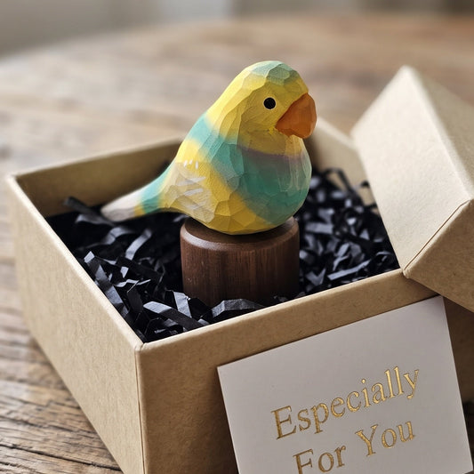 A small hand-painted wooden Lovebirds figurine in yellow, green, and orange, displayed in a brown gift box with black shredded paper and a card that reads 'Especially For You' on a rustic wooden table.