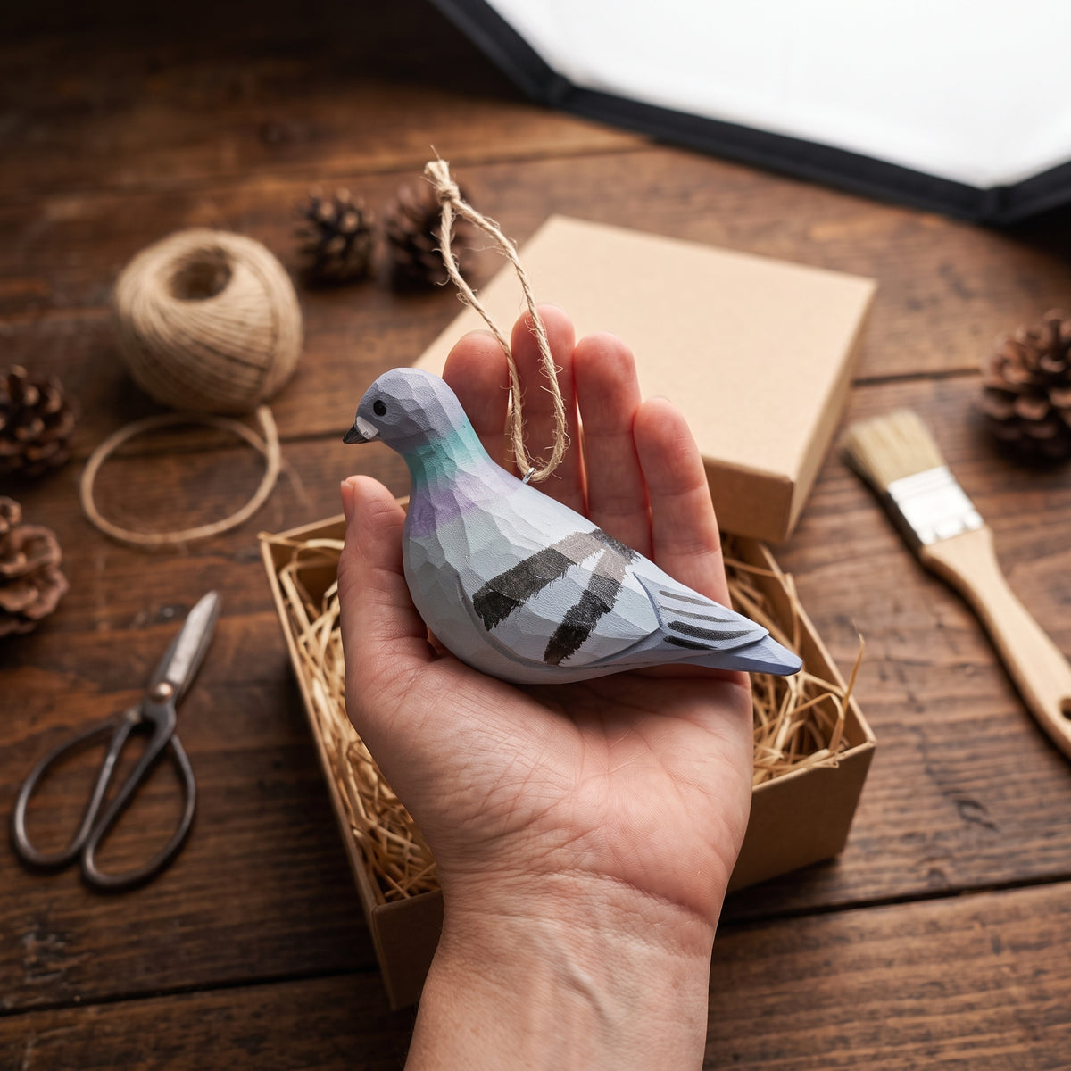 A hand holding a hand-carved and hand-painted wooden Rock Dove pigeon ornament over a rustic craft workspace with a gift box, twine, and scissors.