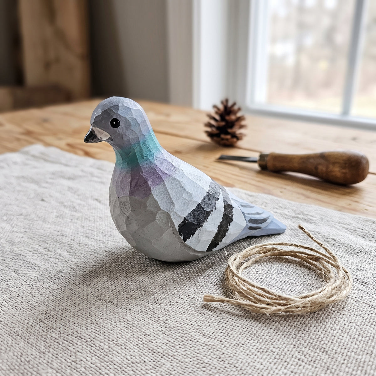 Hand-carved and hand-painted wooden Rock Dove pigeon ornament resting on a coarse linen cloth beside a small coil of twine string and woodcarving tools.