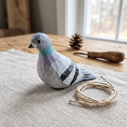 Hand-carved and hand-painted wooden Rock Dove pigeon ornament resting on a coarse linen cloth beside a small coil of twine string and woodcarving tools.