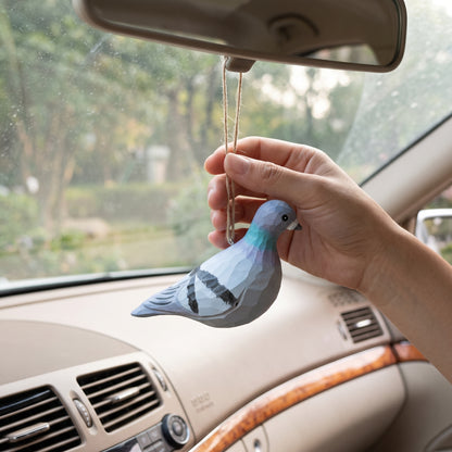 A hand holding a hand-carved and hand-painted wooden Rock Dove pigeon ornament by its twine loop inside a car near the rearview mirror.