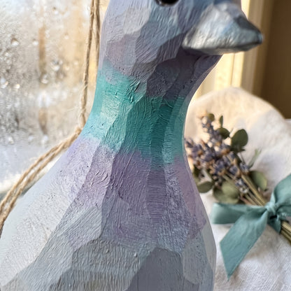 Close-up of the neck and chest of a hand-carved wooden Rock Dove pigeon ornament, showing hand-painted green and purple feather facets with a bouquet of dried lavender in the background.