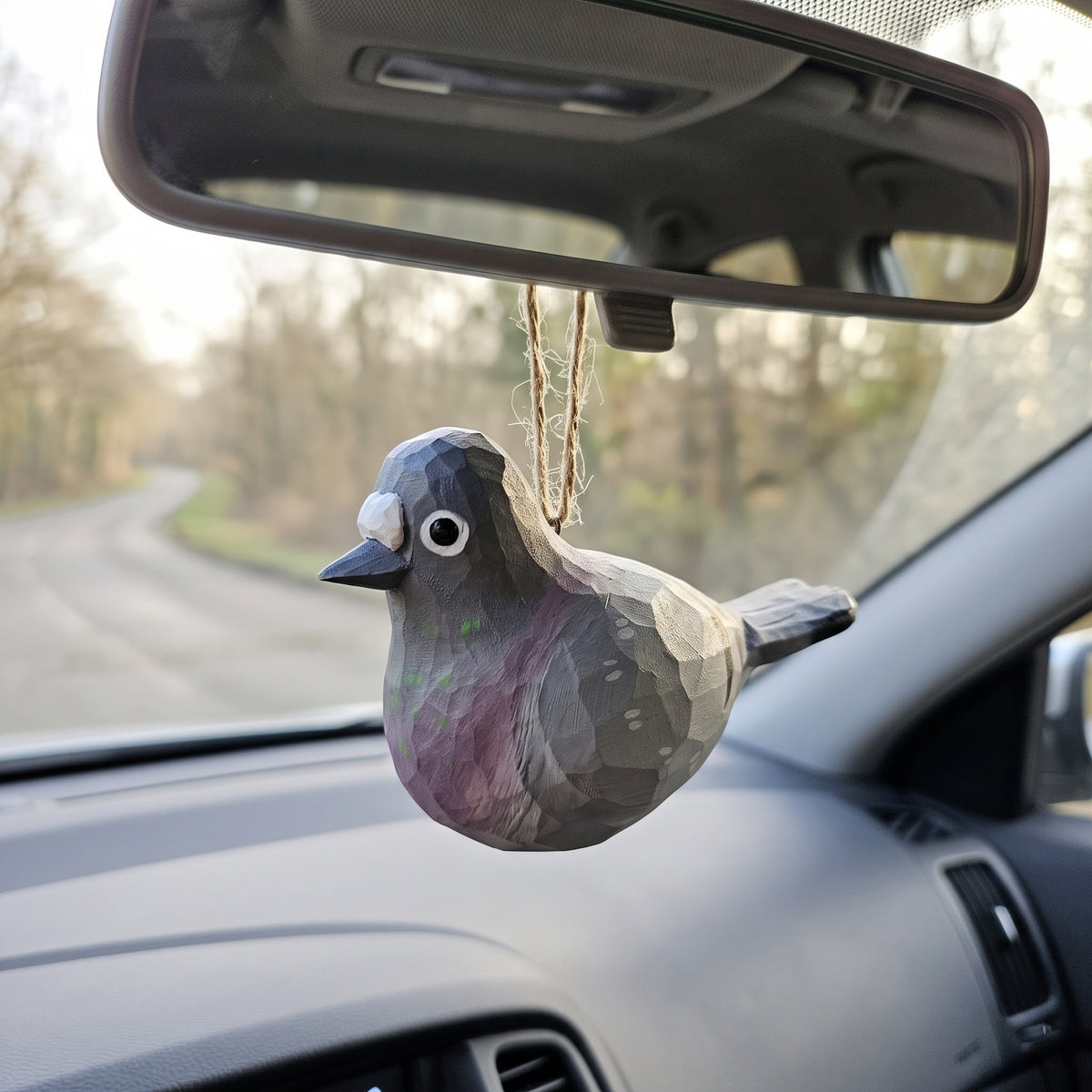 A faceted bird-shaped car air freshener hanging from a car's rearview mirror, with a blurred road and trees visible through the windshield.