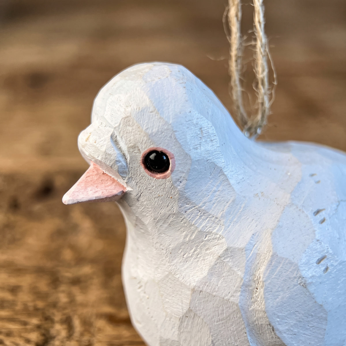 A close-up of a handcrafted wooden pigeon  ornament painted in light blue and white, with a pink beak and a hanging twine loop, set against a warm, blurred wood background.
