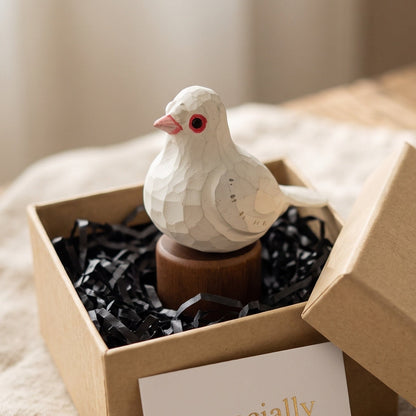 A small white Pigeons figurine with red eyes and a pink beak sits on a wooden base inside an open kraft gift box filled with black paper shred. A card reading 'Especially For You' rests in front, and the box lid leans against the right side on a soft beige fabric surface.