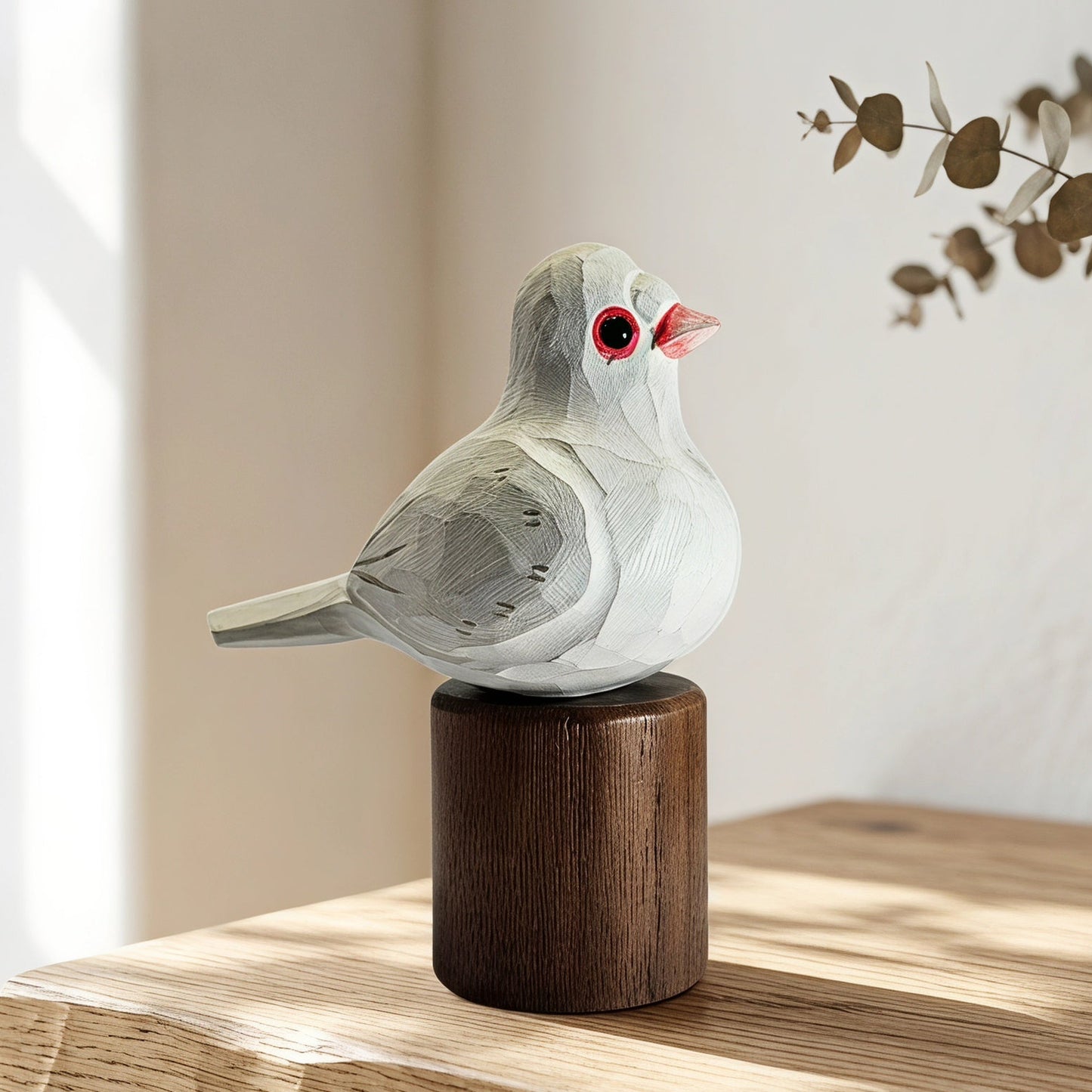 A small carved Pigeons figurine with a white and gray body, red eye, and pink beak, mounted on a dark brown cylindrical wooden base and placed on a light wood table in a softly lit interior setting.