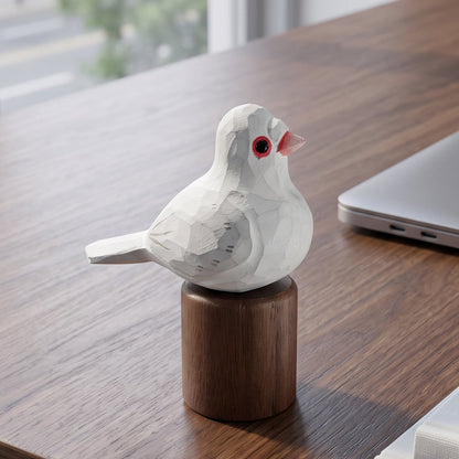 A small white Pigeons figurine with a red eye and pink beak perched on a round wooden base, placed on a dark wood desk next to a closed laptop, notebooks, and a pen.