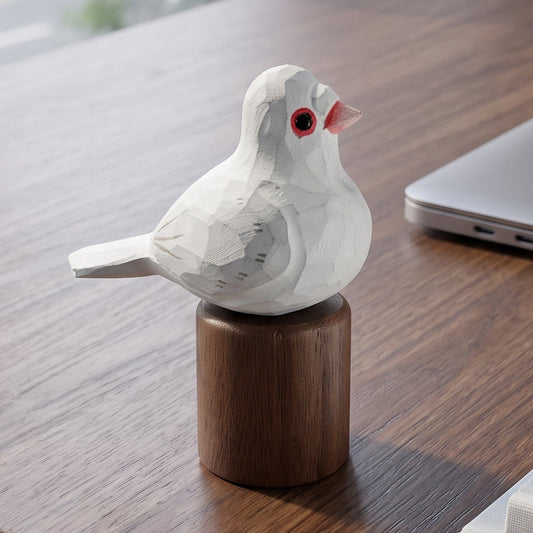 A small white Pigeons figurine with a red eye and pink beak perched on a round wooden base, placed on a dark wood desk next to a closed laptop, notebooks, and a pen.