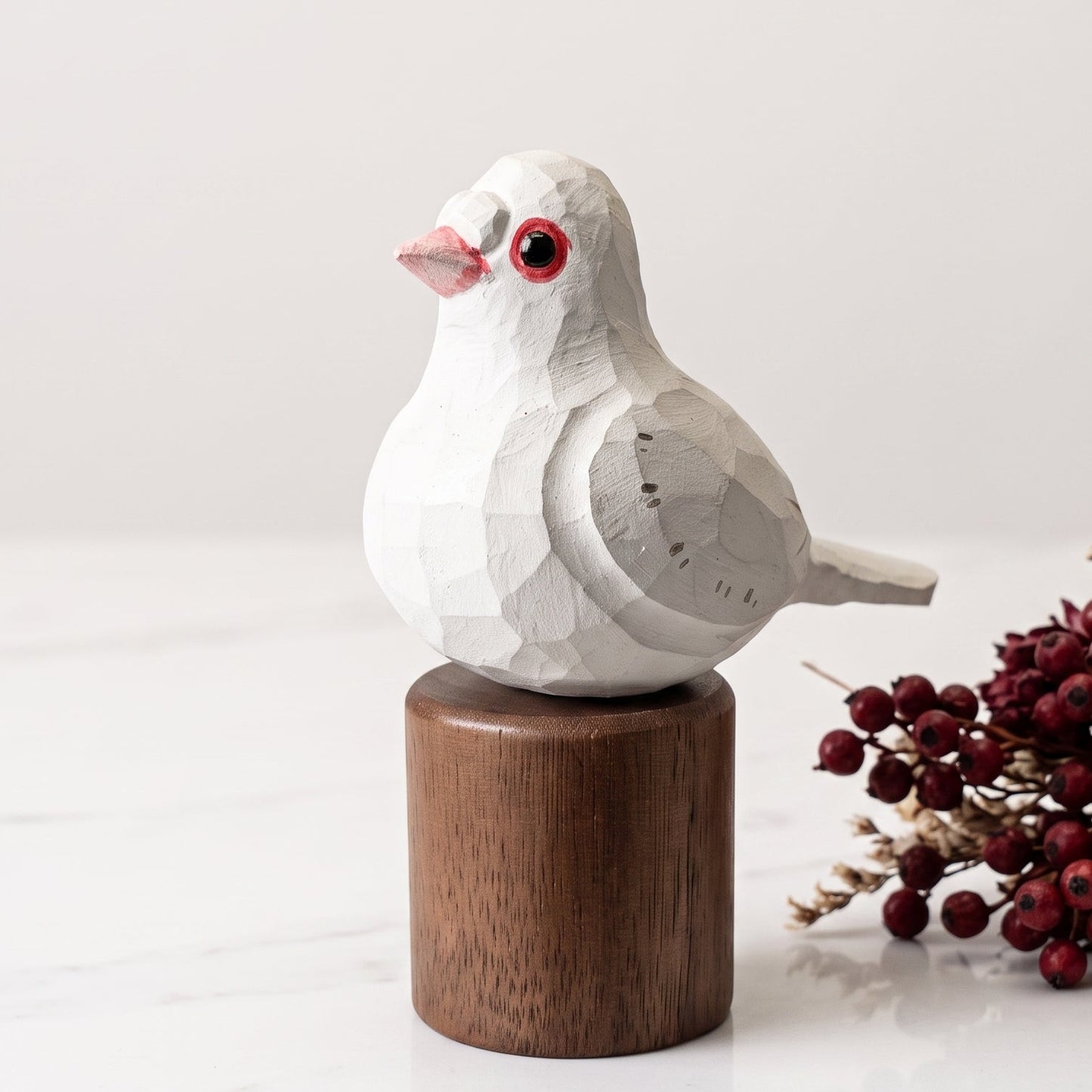 A small white Pigeons geometric bird figurine with a pink beak and red eye, mounted on a round brown wooden base, set on a white marble surface with dried red berries and beige flowers beside it.