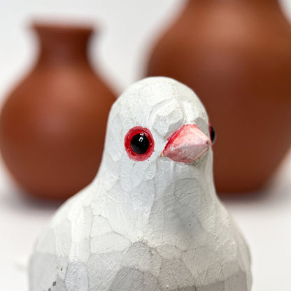 A close-up of a white textured Pigeons figurine with red-pink beak and red-rimmed eyes, with two blurred brown ceramic vases in the background.