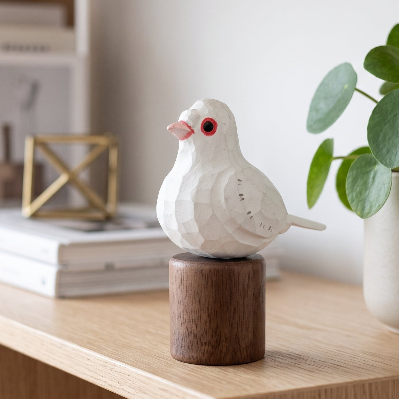 A white geometric Pigeons figurine with a pink beak and red eye, mounted on a dark brown wooden cylinder, displayed on a light wood shelf next to a potted plant.