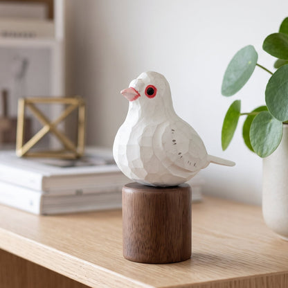 A white geometric Pigeons figurine with a pink beak and red eye, mounted on a dark brown wooden cylinder, displayed on a light wood shelf next to a potted plant.