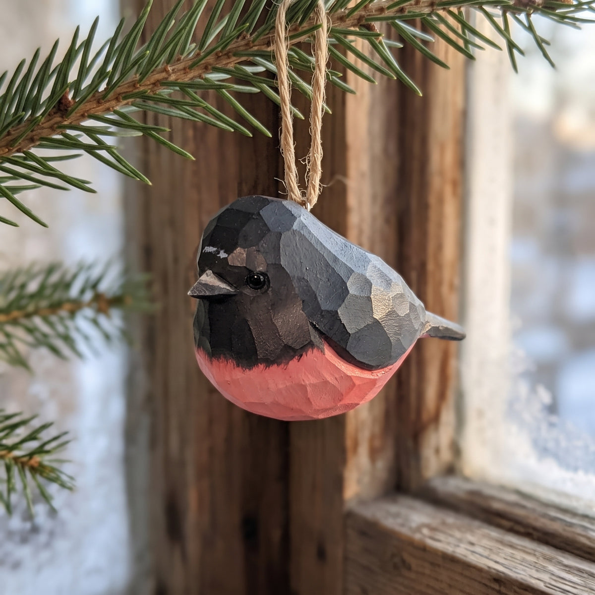Hand-carved wooden pink robin ornament hanging from a pine branch by a snowy window.