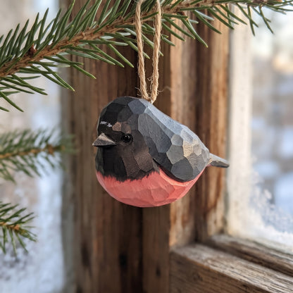 Hand-carved wooden pink robin ornament hanging from a pine branch by a snowy window.