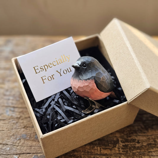 A hand-carved wooden Pink Robin clip-on ornament resting inside an open kraft gift box filled with black shredded paper, featuring a dark slate-grey crown and back, a vivid rose-pink breast, a dark eye, a metal clip underneath, and a white card reading "Especially For You" placed in the foreground on a rustic wooden surface.